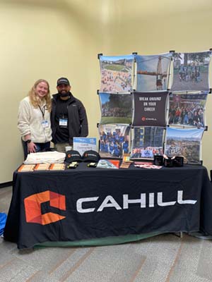 Two people standing behind a table with a sign that says "Cahill"