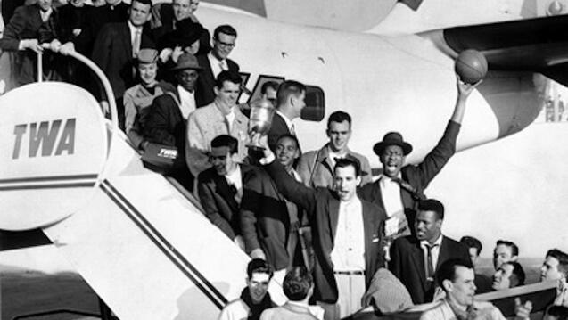 1955 Dons Basketball team walk down stairs leaving a plane, with Bill Russell holding up a game basketball