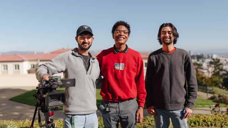 Three students standing next to a film camera