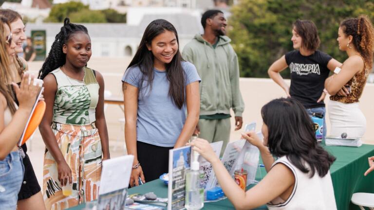 4 students standing and listening to representative seated at a tabling event.