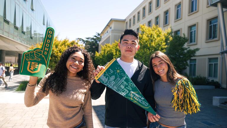 Three students posing on campus waving USF merch