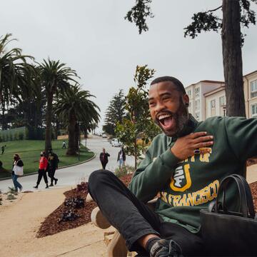 Student in USF sweatshirt sits on bench in front of Lone Mountain dorms.