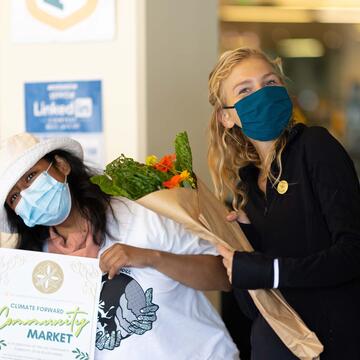 Two students pose for a picture, one holding flowers and the other a sign that reads “Climate Forward Community Market”