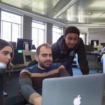 Three students huddled around a laptop in computer lab