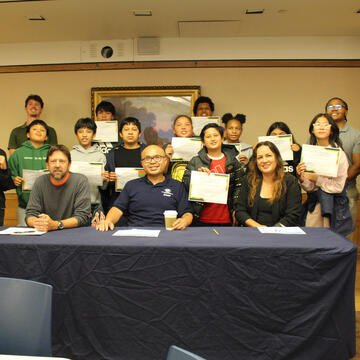 Three representatives sitting at a table with kids behind them holding certificates