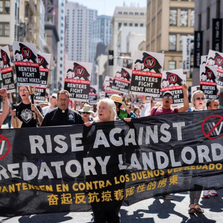 Protestors march carrying a banner that says "Rise Against Predatory Landlords"